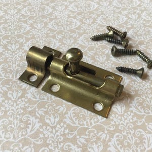 May include: A brass-colored slide bolt latch with a round knob and several screws. The latch is rectangular with rounded edges and circular holes for mounting. The screws are dark metal. The items are on a patterned surface.
