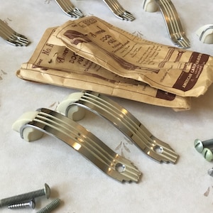 Op de afbeelding: Een close-up van een set vintage chromen en witte kastdeurgrepen met een verpakkingsetiket met de tekst "American Cabinet Hardware, Philadelphia, Pa."