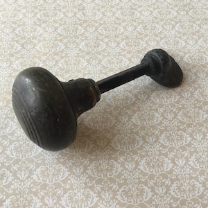 Peut inclure: Bouton de porte ancien avec une finition sombre et vieillie. Le bouton a une forme arrondie avec des rainures verticales, relié par une tige noire à un bouton plus petit et assorti. Une pièce de quincaillerie vintage.