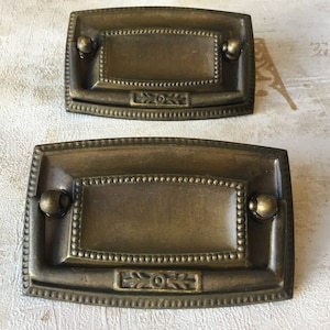 Op de afbeelding: Twee antieke messing lade grepen. Elke rechthoekige greep heeft een centraal verzonken paneel, een kralenrand en afgeronde knoppen. De grepen hebben een decoratief bloemmotief in het midden.