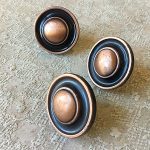 May include: Three round copper cabinet knobs with black trim. The knobs have a raised centre and a slightly recessed outer ring.
