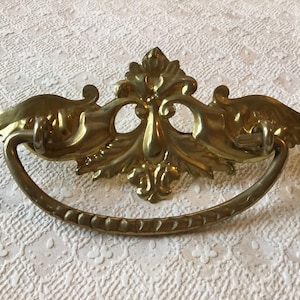 May include: A gold-toned ornate brass drawer pull with a decorative, scrolled design. The pull has a curved, D-shaped handle.