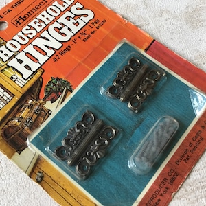 May include: A pack of two decorative brass hinges, each measuring 2.5 cm by 1.9 cm. The pack is labelled "Household Hinges #2 Hinge - 1" x 3/4" - 1 Pair Stock No. 657220" and "EPRODUCER CO. Division of Goats & Clark Inc. New York 10802 Pat. Pending Made in U.S.A."
