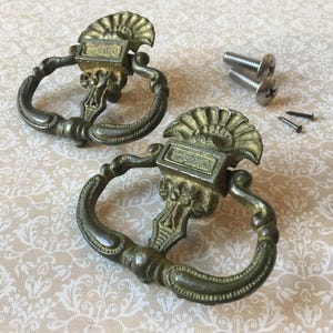 May include: Two antique brass drawer pulls with ornate detailing and a fan-shaped top. The pulls have a curved handle and are accompanied by several screws for installation. A vintage hardware item.