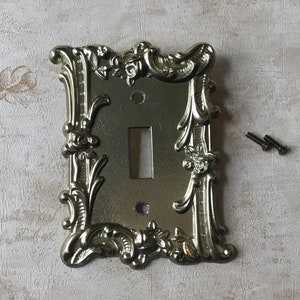 May include: Gold-toned decorative light switch cover with an ornate, floral design. The cover has a single switch opening and comes with two screws.