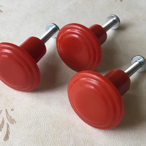 Puede incluir: Tres tiradores de armario rojos con un diseño redondeado y acanalado. Cada tirador tiene un tornillo plateado. Los tiradores están dispuestos sobre una superficie clara con un patrón sutil.