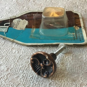 May include: A bronze cabinet knob with a decorative floral design and a silver screw. The knob is packaged in a clear plastic bag with a blue and white label that says "Eaton's".