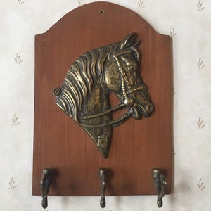 Peut inclure: Un porte-clés en bois avec un motif de tête de cheval en laiton. La tête de cheval est détaillée avec une bride et un mors. Il y a trois crochets au bas du porte-clés.