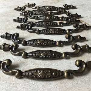 May include: Ten antique brass cabinet door pulls with ornate floral designs. Each pull has a curved handle and a decorative knob.