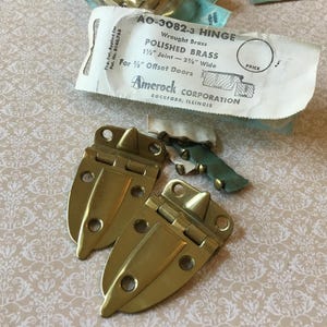 Puede incluir: Dos bisagras de latón pulido con una etiqueta de papel que dice "AO-3082-3 HINGE" y "POLISHED BRASS". La etiqueta también indica que la bisagra es para puertas desplazadas de 9,5 mm. Las bisagras son de color dorado y están sobre un fondo estampado.
