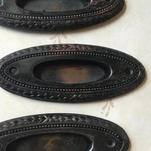 Puede incluir: Tres tiradores de cajón antiguos, de forma ovalada y con un acabado oscuro y envejecido. Cada tirador tiene un centro empotrado y está enmarcado por un borde detallado con cuentas y detalles florales decorativos. Los tiradores son probablemente de metal.