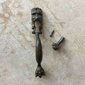 May include: Antique bronze cabinet pull with a detailed face design and claw-foot base. Includes two detached screws. The hardware is likely for a drawer or cabinet.