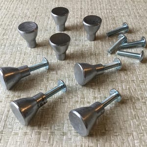 May include: A selection of silver cabinet knobs and screws. The knobs have a rounded top and a conical base. The screws are silver and have a slotted head. The hardware is arranged on a textured, light-coloured surface.