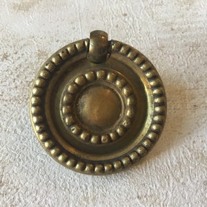 May include: A vintage brass drawer pull with a circular design. The pull features a central disc surrounded by two concentric rings, one with a beaded texture. A small, arched handle is attached at the top. The hardware has a warm, aged patina.