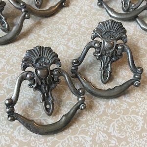 May include: A selection of antique silver-toned drawer pulls. Each pull has an ornate design with a central floral motif and a curved handle. The pulls are displayed on a cream-coloured surface with a floral pattern.
