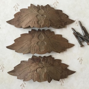 Puede incluir: Tres tiradores de cajón antiguos, marrones, con forma de hoja y diseño de ojo de cerradura. Los tiradores son de madera oscura y van acompañados de un pequeño montón de tornillos. El fondo es claro y estampado.