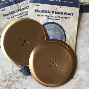 May include: Two gold-coloured metal back plates, each with five holes. The plates are labelled "No. E502-10 BACK PLATE" and are for use with concave knobs.