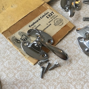 May include: Chrome-plated cupboard catches and screws are arranged on a patterned beige surface. A vintage cardboard box, labelled "Cupboard Catches 1327", is partially visible. The hardware is designed for cabinets.