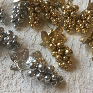 May include: An assortment of decorative grape clusters in silver and gold. Each cluster features small, round beads representing grapes, attached to metallic leaves and stems. The gold clusters have a shiny finish, while the silver ones have a matte appearance.