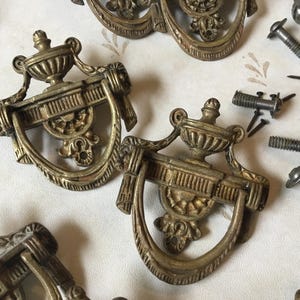 Puede incluir: Primer plano de tiradores de cajón antiguos de latón con detalles ornamentados, incluido un diseño central de urna y asas curvas. Varios tornillos están esparcidos alrededor de los tiradores, lo que sugiere que están listos para la instalación. El hardware tiene una estética vintage.