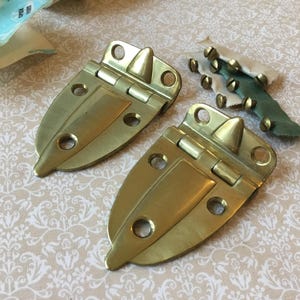 Peut inclure: Deux charnières dorées avec un design pointu, chacune avec trois trous pour les vis. Une petite pile de vis en laiton et un morceau de tissu vert sont également visibles. Les charnières sont probablement pour une armoire ou une petite porte.