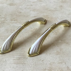 May include: Two cabinet handles with a white center and gold-colored trim. The handles have a curved design with a textured gold accent near the top. The handles are approximately 5 inches long.