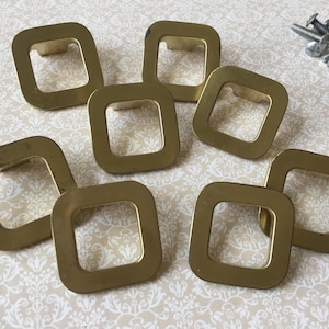 May include: A collection of seven square brass cabinet pulls with rounded corners. The pulls are arranged on a patterned surface, with a small pile of silver screws in the upper right corner. The pulls have a slightly aged appearance.
