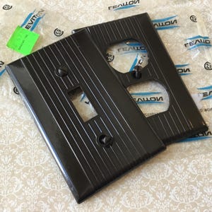 Pode incluir: Duas placas pretas para tomadas e interruptores com linhas verticais. As placas são novas e ainda estão na embalagem original. A embalagem tem a marca "Leviton" impressa.