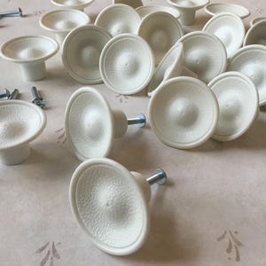 Op de afbeelding: Een verzameling witte keramische kastknoppen met een textuur. De knoppen hebben een eenvoudig, rond ontwerp en een klein schroefgat in het midden.
