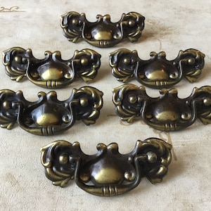 May include: Six antique brass cabinet pulls with ornate floral designs. Each pull has a rounded, slightly raised centre with a small loop handle.