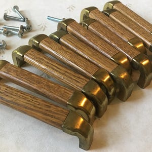 May include: A set of eight wooden cabinet handles with brass-coloured metal detailing. The handles have a rectangular shape with rounded corners, and are shown with a collection of silver screws.