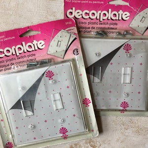 May include: Two packages of clear plastic light switch plates with a decorative seashell design. The packages are pink and white and include screws for installation. The text on the package reads "decorplate clear plastic switch plate plaque de commutateur en plastique transparent SP-02"