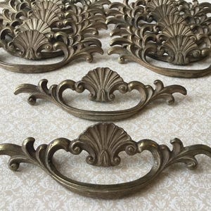 Peut inclure: Plusieurs poignées de tiroir antiques en laiton avec un motif de coquillage et de volutes. Les poignées ont une poignée incurvée et des détails ornés. La quincaillerie est de couleur or mat et est probablement en métal.