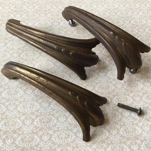May include: Three antique bronze-coloured drawer pulls with a decorative, curved design. The pulls have a detailed, textured surface and are accompanied by a single screw. The hardware is likely for a cabinet or dresser.