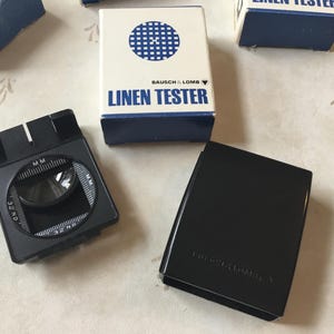 May include: A black linen tester with a magnifying glass and a dial marked in millimetres. The tester is made by Bausch & Lomb. The box for the tester is white with blue trim and says "Linen Tester" in bold blue letters.