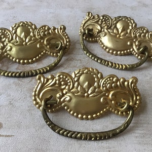 Op de afbeelding: Drie sierlijke, goudkleurige ladegrepen met een decoratief, gerold ontwerp. Elke greep heeft een gebogen, lusvormige handgreep.