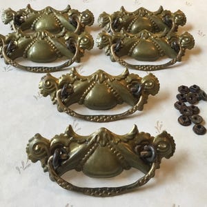 May include: Six antique brass drawer pulls with ornate designs and a set of screws. The pulls have a decorative, ornate design with a heart-shaped center.