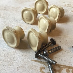 Peut inclure: Six boutons de porte en céramique blanc cassé avec des bords arrondis et un centre surélevé. Les boutons sont disposés en rangée avec des vis éparpillées autour d'eux.
