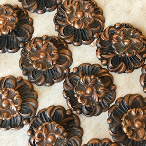 Puede incluir: Un conjunto de 12 adornos metálicos de color cobre con un diseño floral. Cada adorno tiene un centro elevado con un diseño circular y un fondo texturizado negro.