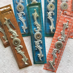 May include: A close-up of eight packages of decorative drawer pulls. The pulls are made of metal and have a white finish with gold accents. The packages are labeled "Amerock" and have a variety of designs.