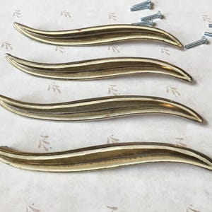 May include: Four gold-toned, curved cabinet handles with a central white stripe. The handles are arranged horizontally, with several silver screws scattered nearby. The handles have a sleek, modern design, suitable for furniture restoration or new builds.