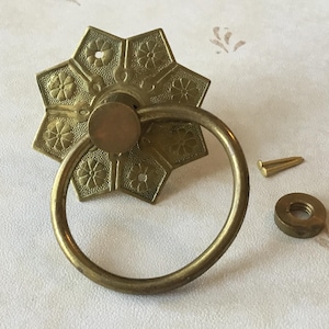 Könnte beinhalten: Vintage Schubladengriff aus Messing mit Sternmuster und rundem Griff. Enthält eine Schraube und eine Mutter. Die Hardware hat eine warme, gealterte Patina, was darauf hindeutet, dass es sich um ein antikes oder Vintage-Stück handelt.