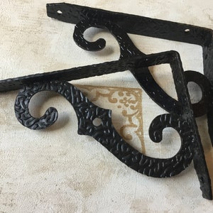 Puede incluir: Dos soportes de estante de metal negro con un diseño decorativo de desplazamiento. Los soportes tienen un acabado texturizado y están en foco.