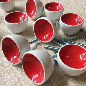May include: A set of white ceramic cabinet knobs with a bright red interior. Each knob is a rounded bowl shape, and several have silver screws attached. The knobs are arranged on a patterned surface.
