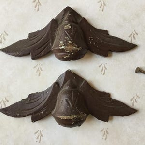May include: Two antique, brown metal corner brackets with a decorative, winged design. The brackets have a distressed finish, showing signs of wear. Several screws are visible nearby. These are likely for furniture restoration or decorative projects.