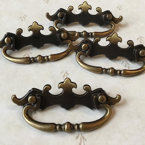 Peut inclure: Ensemble de quatre poignées de tiroir en laiton antique avec des motifs ornés. Les poignées ont une finition noire et une poignée incurvée en forme de D.