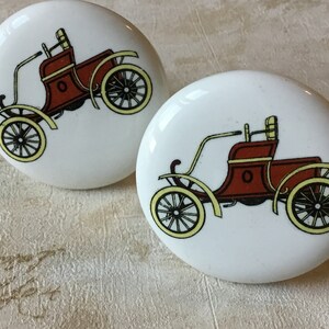 Può includere: Due pomelli in ceramica bianca con un design di auto d'epoca rossa. Le auto hanno ruote gialle e dettagli neri.