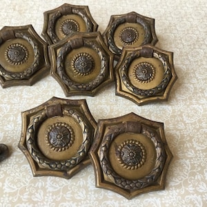Puede incluir: Colección de seis tiradores de puerta antiguos de latón. Cada tirador presenta un diseño circular central con un motivo floral, rodeado por una guirnalda decorativa y enmarcado en un marco octogonal. Aspecto envejecido.