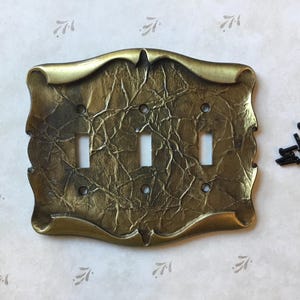 May include: A decorative triple light switch cover in a metallic gold finish. The cover has a textured, wrinkled surface and an ornate, curved design. Black screws are included.