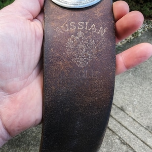 Puede incluir: Una correa de afeitar de cuero marrón con una hebilla de metal plateado y la palabra "RUSSIAN" grabada en el cuero. El cuero tiene un aspecto desgastado y envejecido.